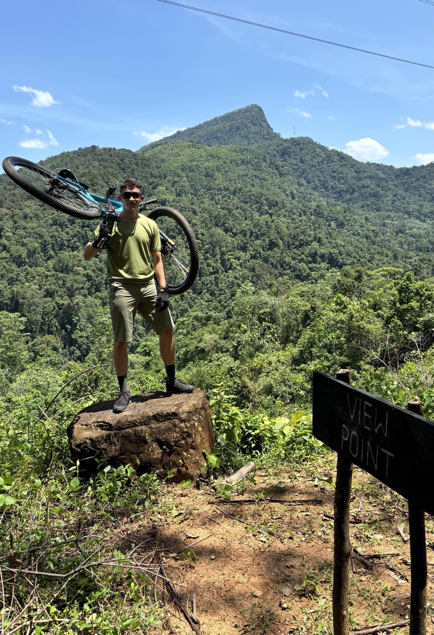 A rider stands on a rock and lifts his bike over his shoulder, with a forested peak behind him